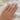 Hand wearing a two ring set; one round diamond solitaire ring and the other, a thin, curved gold band on a white fabric background.