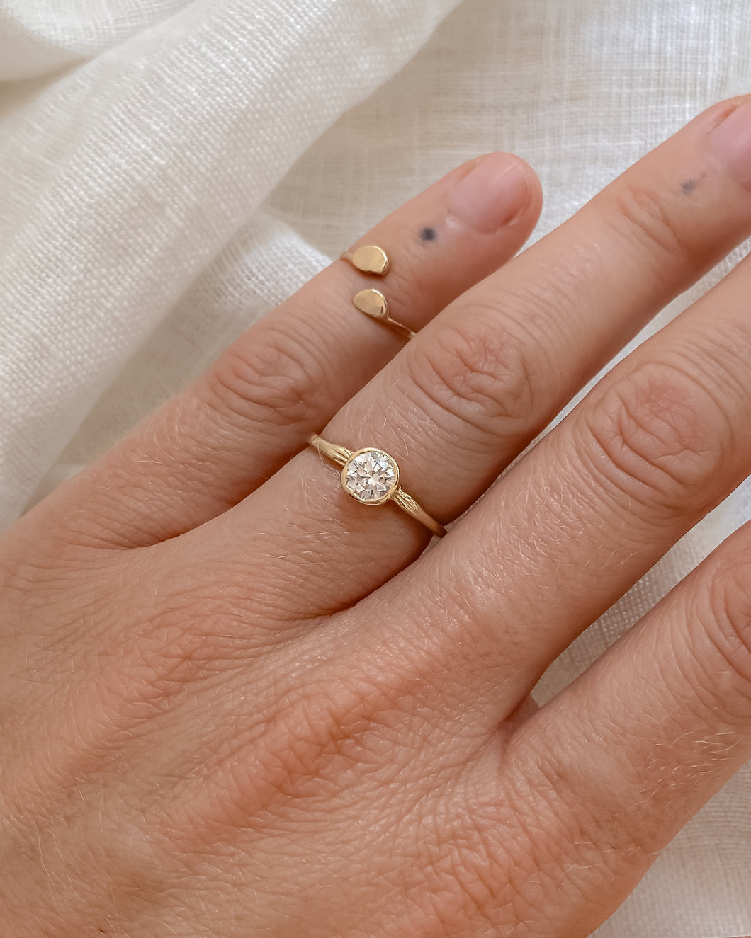 Gold ring with a round diamond in a bezel setting, on a hand against a light fabric background