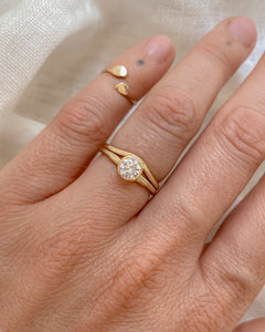 A round diamond, solitaire, gold ring on a hand against a neutral background. The solitaire is paired with a slightly curved, thin gold band.