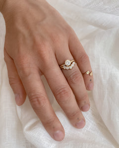 Hand wearing two diamond rings on a white fabric background; one ring is a round, diamond solitaire and the other is a curved, diamond half halo ring.