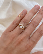 Load image into Gallery viewer, Hand wearing a gold, solitaire ring paired with a curved, five diamond band on a white fabric background
