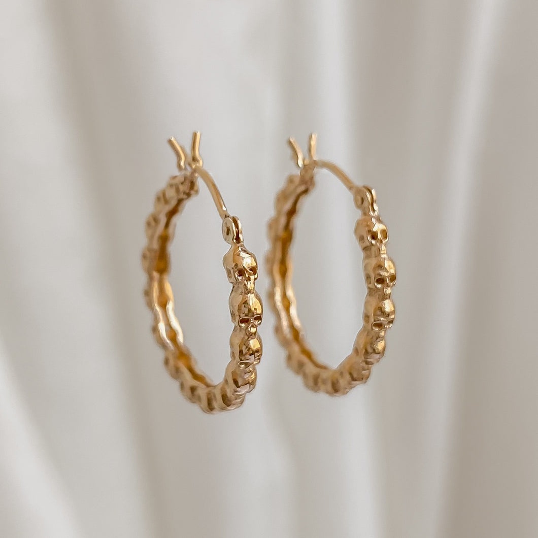 A pair of gold hoop earrings with hand-carved skull designs, displayed against a white fabric background.
