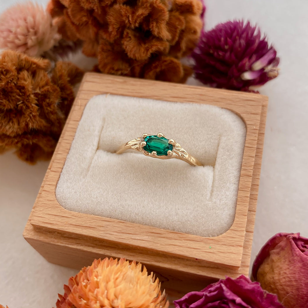 A vintage emerald ring set in a recycled 14k gold band, displayed in a wooden ring box with a white interior, surrounded by colorful flowers.