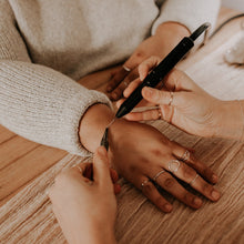 Load image into Gallery viewer, A person is getting a permanent jewelry application on their wrist, held by another person, in a store setting.
