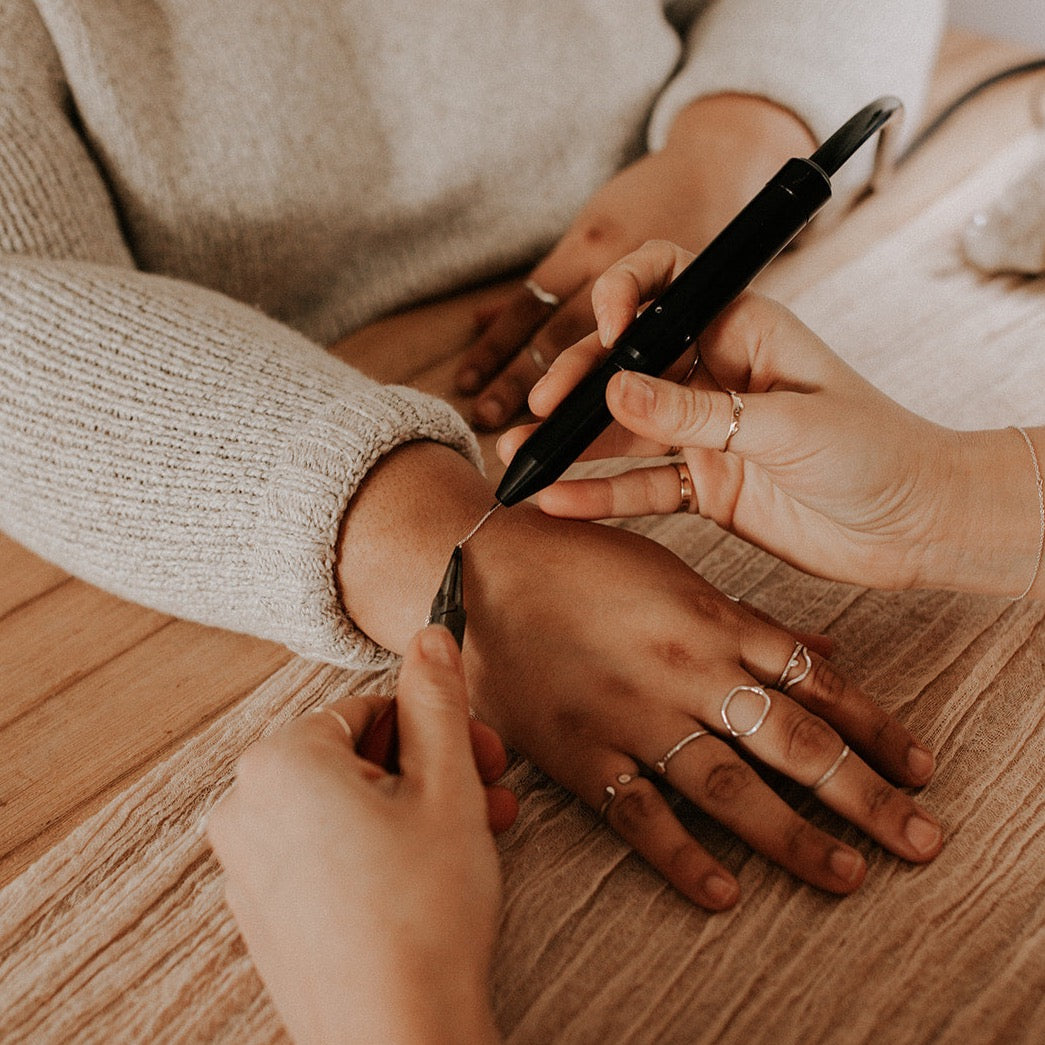 A person is getting a permanent jewelry application on their wrist, held by another person, in a store setting.