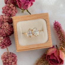 Load image into Gallery viewer, A 14k gold ring with a pear and an oval lab diamond set side by side, presented in a wooden box surrounded by pink flowers.