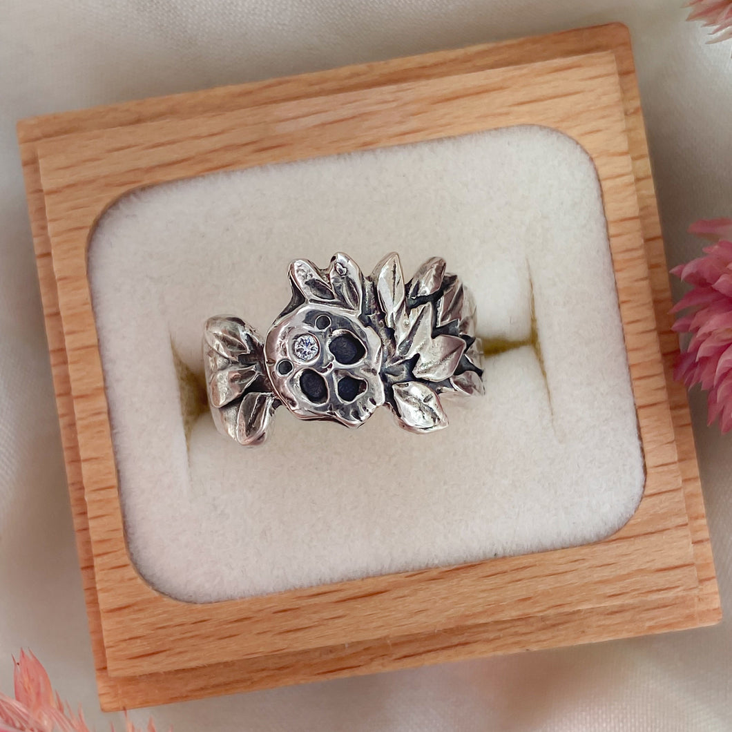 A recycled sterling silver ring with a floral and skull design, set in a ring box on a white cushion.