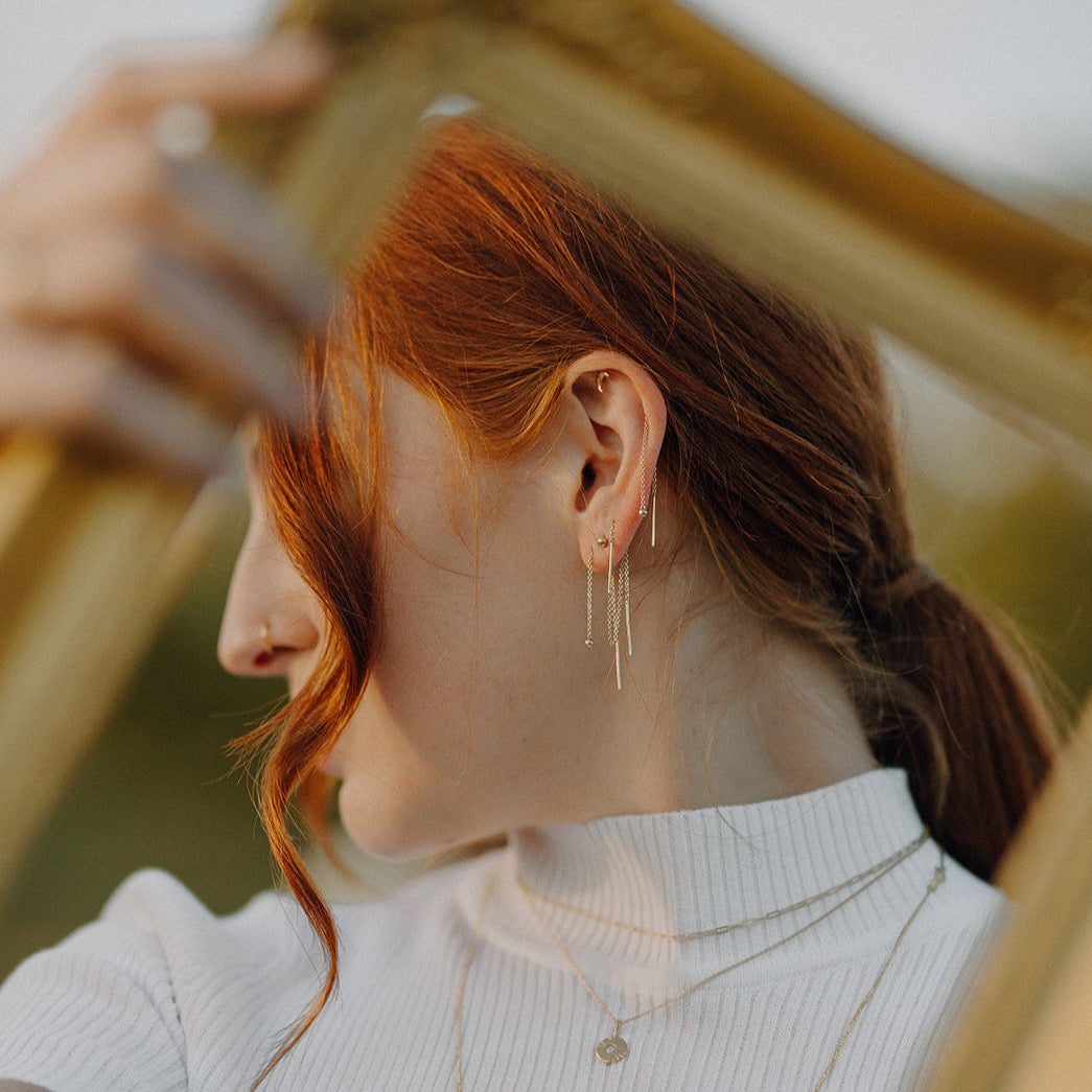 A woman with red hair is seen from the side, wearing small, dangle earrings with a gold chain design.