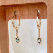 Load image into Gallery viewer, A pair of 14k gold hoop earrings with bi-colored sapphires attached to a wooden display stand.