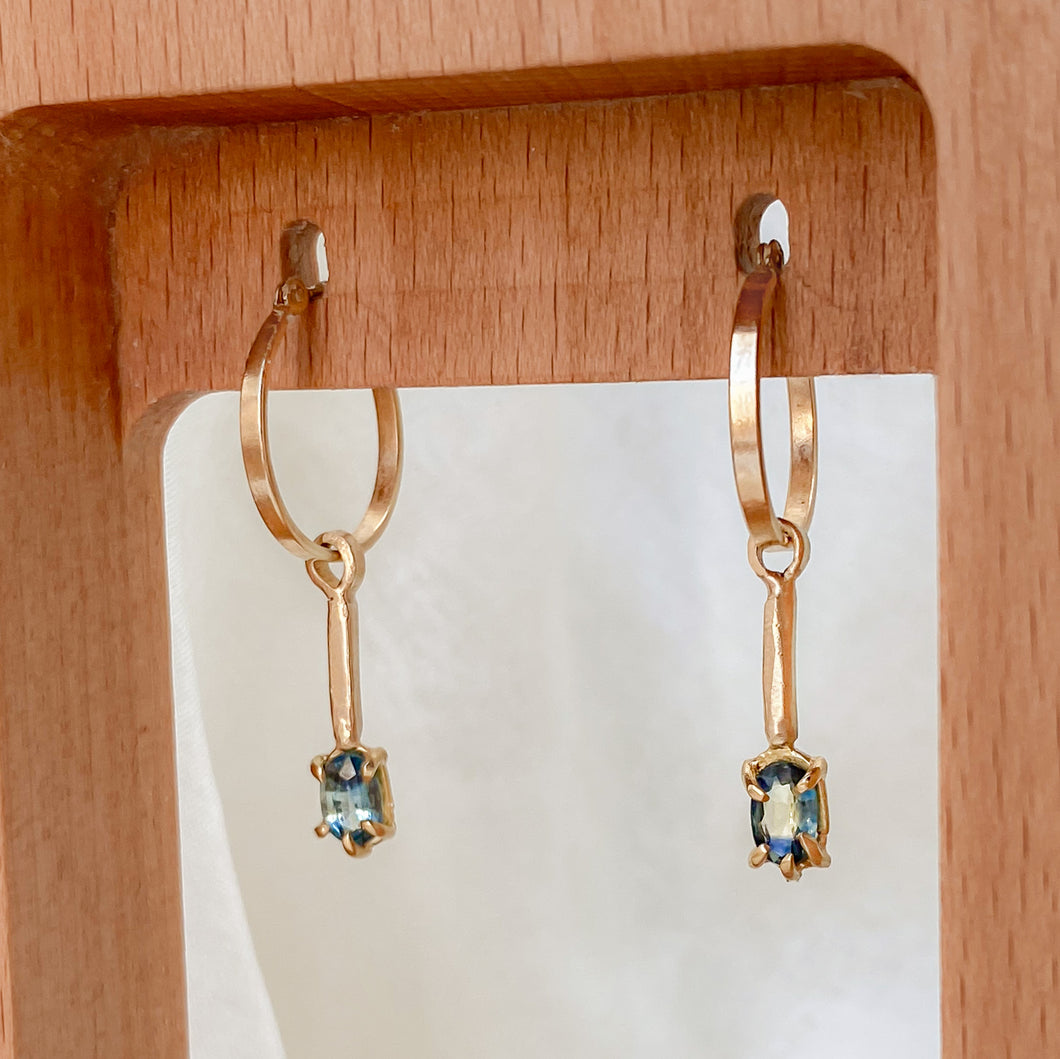 A pair of 14k gold hoop earrings with bi-colored sapphires attached to a wooden display stand.
