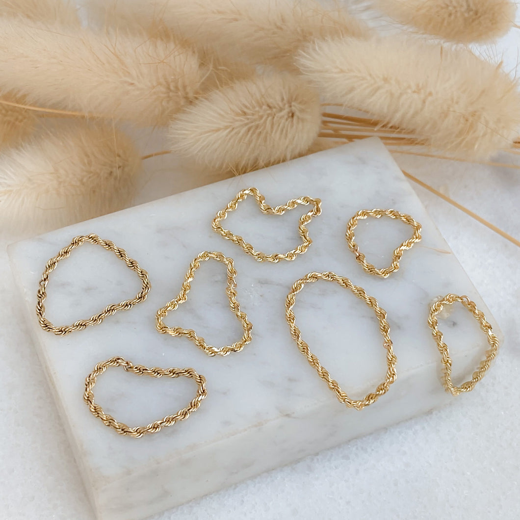 A collection of gold rope chain rings displayed on a marble surface with dried plants in the background.